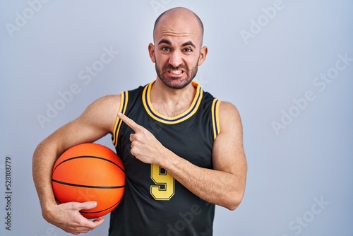 Young bald man with beard wearing basketball uniform holding ball pointing aside worried and nervous with forefinger, concerned and surprised expression