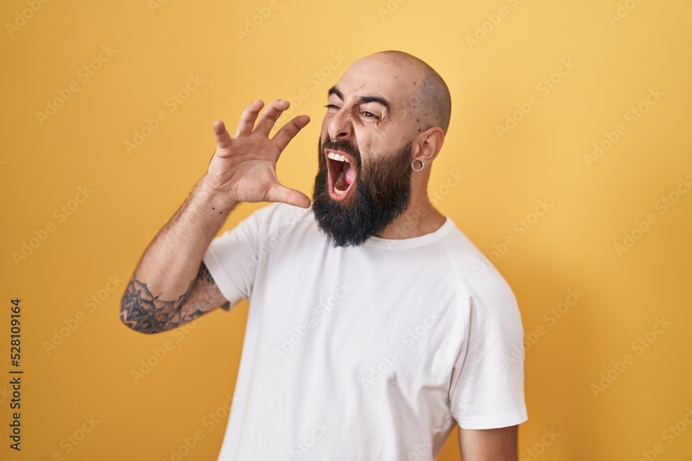 Young hispanic man with beard and tattoos standing over yellow background shouting and screaming loud to side with hand on mouth. communication concept. Young hispanic man with beard and tattoos standing over yellow background shouting and screaming loud to side with hand on mouth. communication concept.