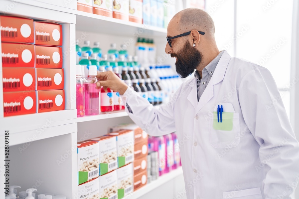 Young bald man pharmacist smiling confident holding medication on shelving at pharmacy