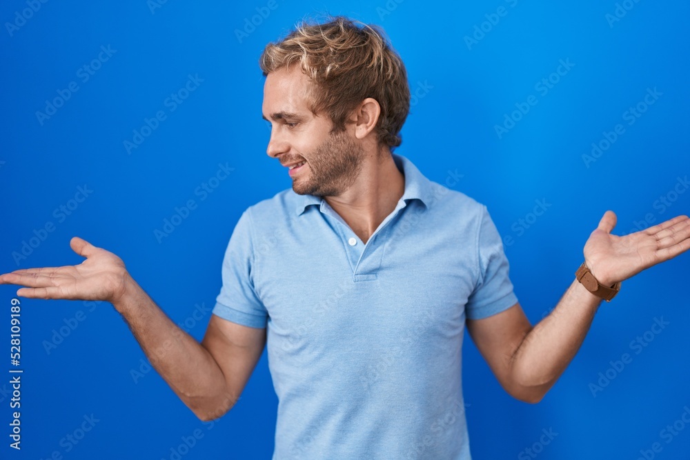Caucasian man standing over blue background smiling showing both hands open palms, presenting and advertising comparison and balance Caucasian man standing over blue background smiling showing both hands open palms, presenting and advertising comparison and balance