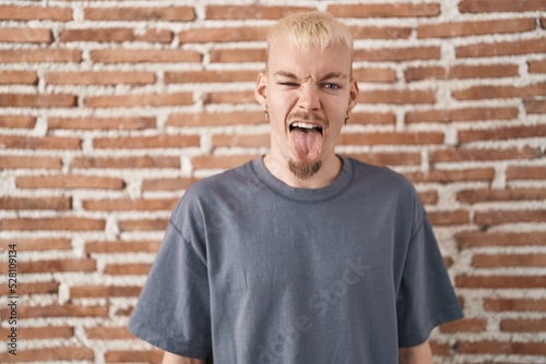 Young caucasian man standing over bricks wall sticking tongue out happy with funny expression. emotion concept.