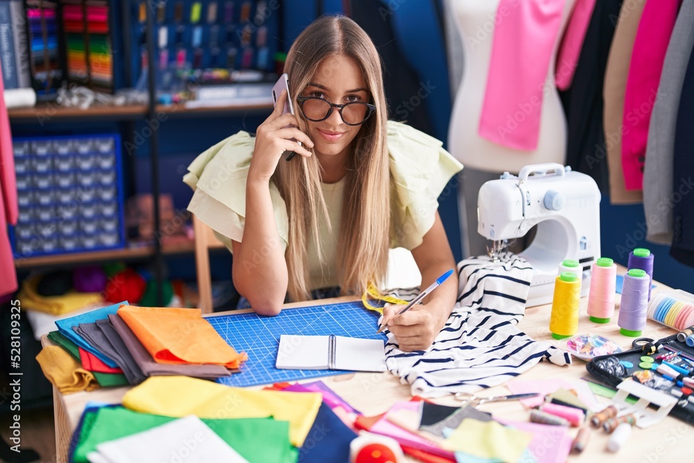 Fototapeta premium Young beautiful hispanic woman tailor talking on smartphone writing on notebook at clothing factory