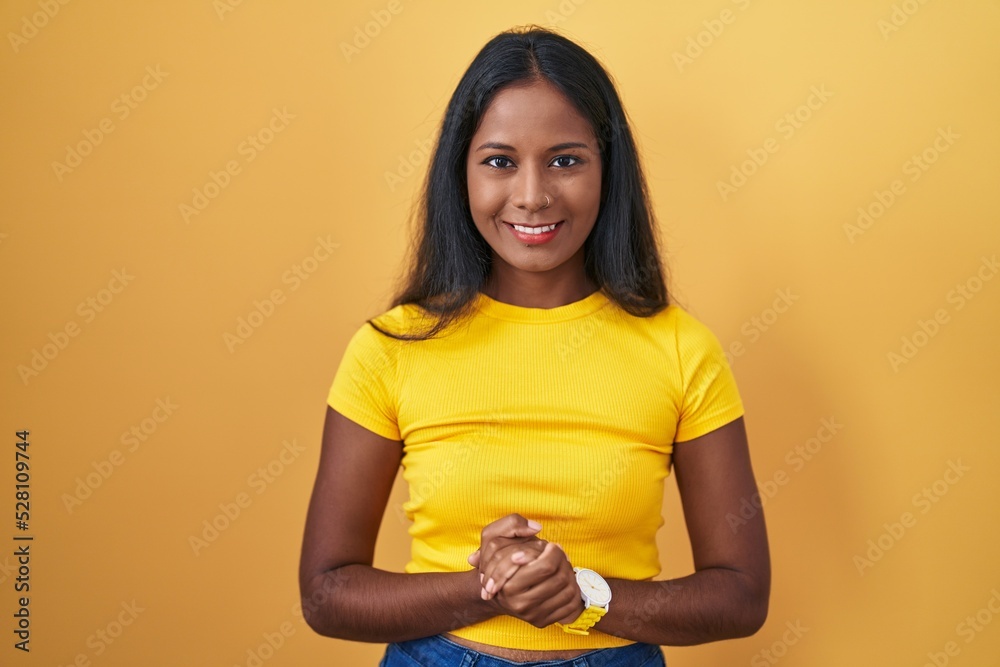 Obraz premium Young indian woman standing over yellow background with hands together and crossed fingers smiling relaxed and cheerful. success and optimistic