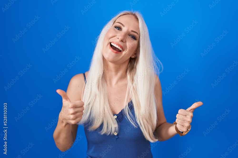 Fototapeta premium Caucasian woman standing over blue background success sign doing positive gesture with hand, thumbs up smiling and happy. cheerful expression and winner gesture.