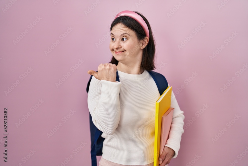 Woman with down syndrome wearing student backpack and holding books smiling with happy face looking and pointing to the side with thumb up. Woman with down syndrome wearing student backpack and holding books smiling with happy face looking and pointing to the side with thumb up.