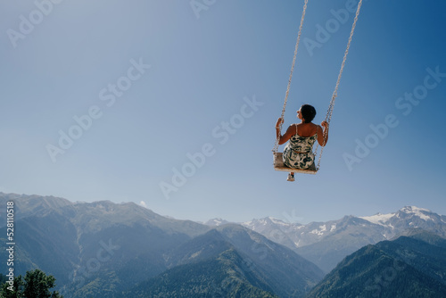 Beautiful woman during summer day flying on swing in blue sky over a mountain landscape. Dream and travel concept