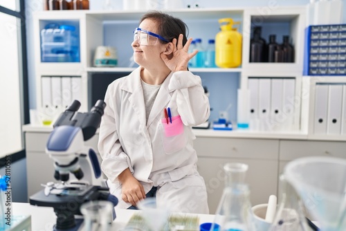 Hispanic girl with down syndrome working at scientist laboratory smiling with hand over ear listening an hearing to rumor or gossip. deafness concept.