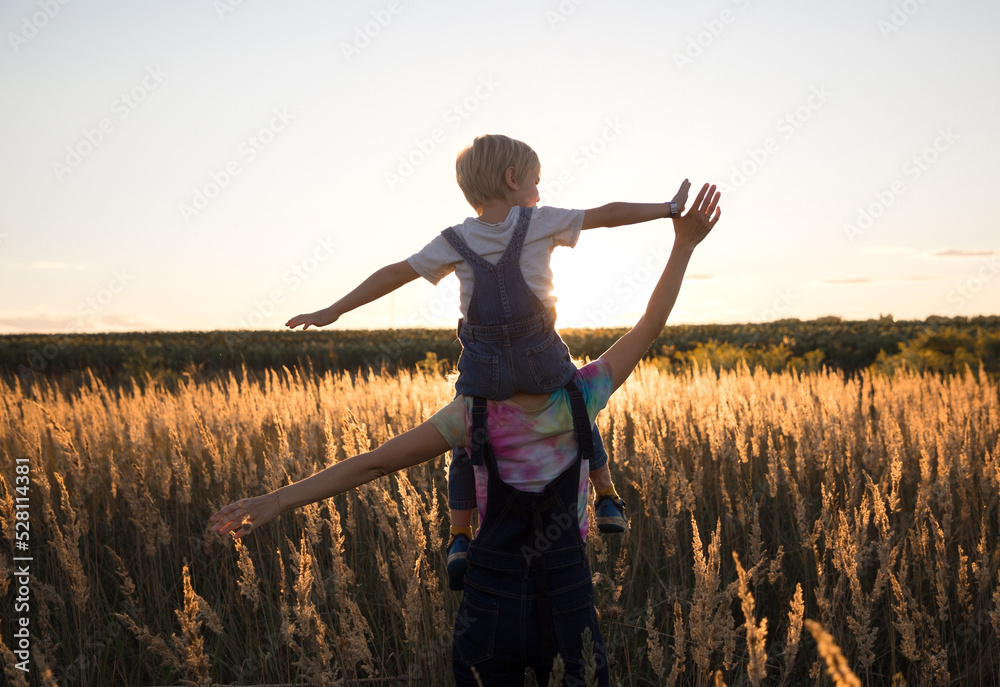 unrecognizable child sits on shoulders of mom and walks along meadow illuminated by sunset light unrecognizable child sits on shoulders of mom and walks along meadow illuminated by sunset light