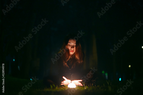 Portrait of beautiful witch in black dress with magic fire ball in forest at dark night