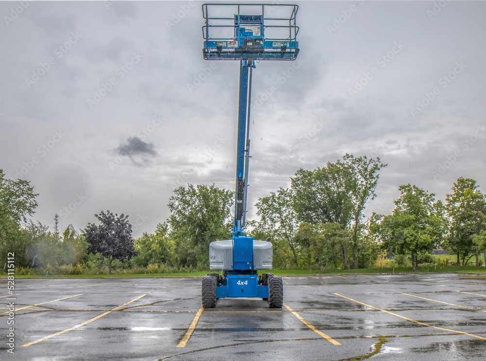 Obraz na płótnie Blue personnel basket lift truck  with hydraulic boom extended upward in an empty parking lot, light rain falling, nobody w salonie