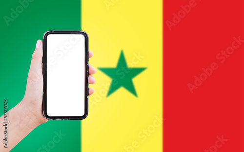 Close-up of male hand holding smartphone with blank on screen, on background of blurred flag of Senegal.