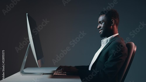 African-American male dark-skinned businessman working at night at a computer monitor in his office against a gray background against a wall