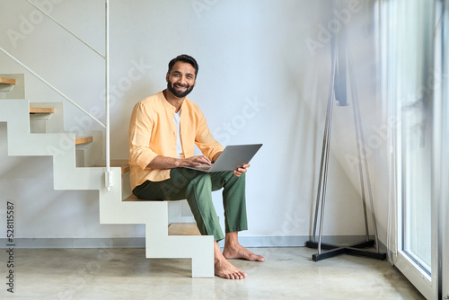 Happy indian man user typing on laptop device, using computer for remote work or elearning, surfing internet digital technology doing online shopping choosing new furniture sitting on stairs at home.