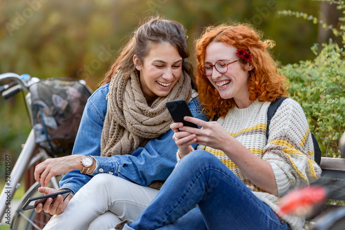 Cheerful female friends sitting on a bench in a park talking, smiling, and using smartphones 