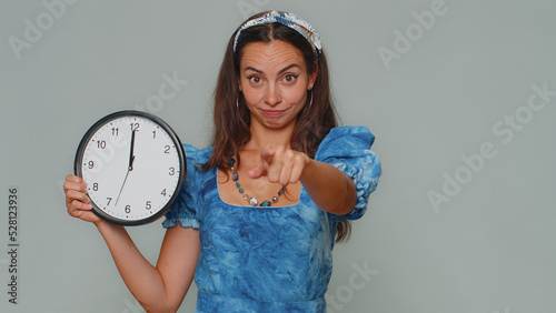 It is your time. Pretty young woman in blue princess dress showing time on wall office clock, ok, thumb up, approve, pointing finger at camera. Stylish female girl on gray studio background indoor photo