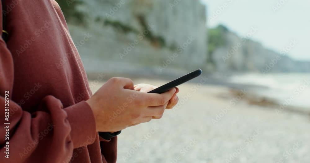 A woman is typing on a smartphone while standing on the seashore near steep limestone cliffs. Close-up of female hands, no face. Etretat, France. 4k video, red komodo