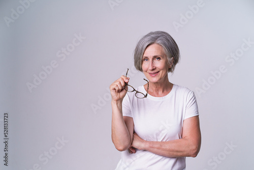 Mature businesswoman took off her glasses with folded hands looking straight in camera and smile wearing white t-shirt isolated on white background. Mature people beauty concept.