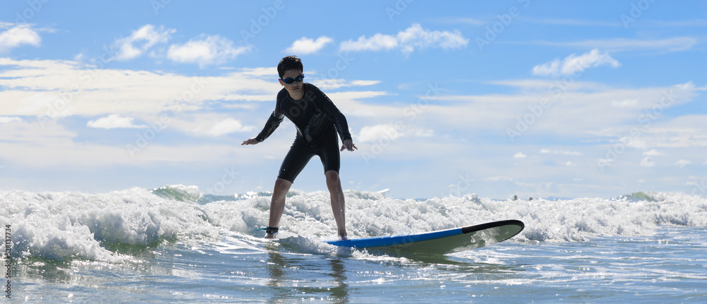 Young boy wearing swimming goggles stable stands on soft board while practicing surfing in beginner's class.