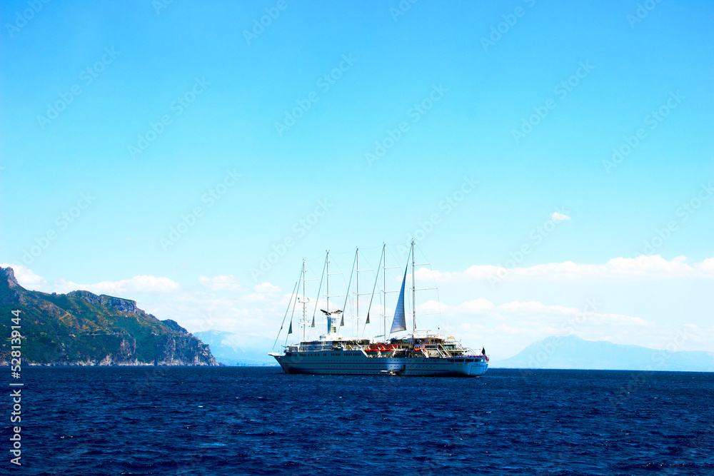 Majestic view in Amalfi coast at a huge colored ship with some sails and emergency vessels skimming the Tyrrhenian Sea with some imposing mountain cliffs in the background under a sunny sky