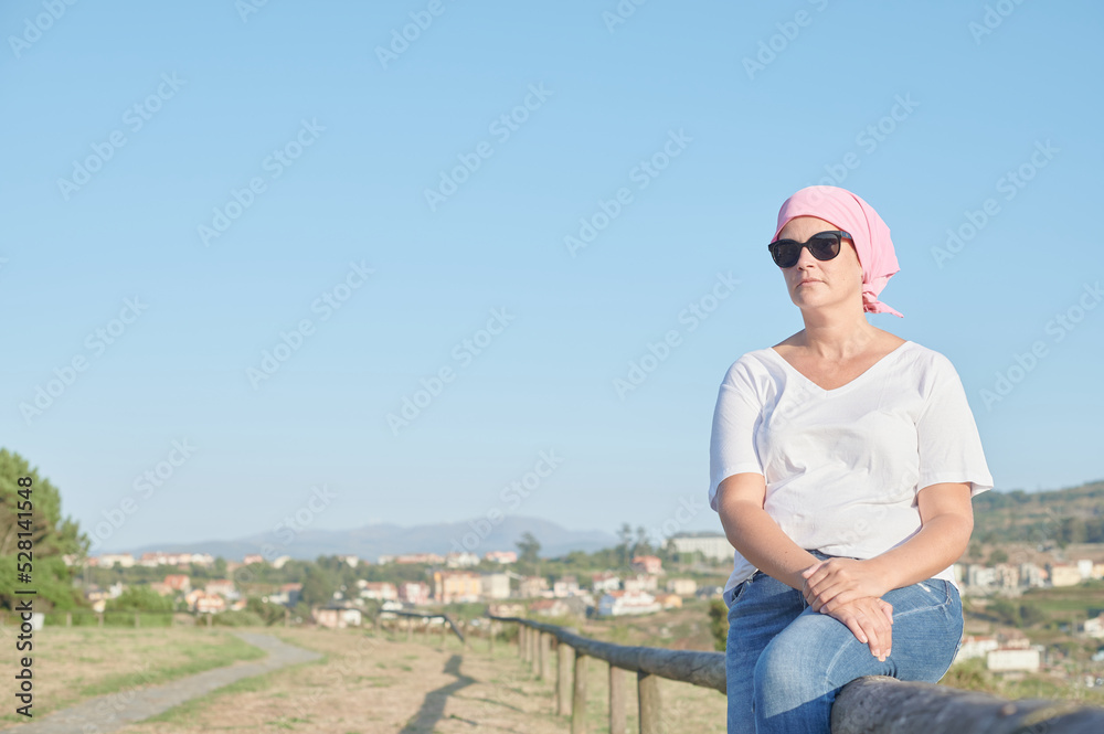 World breast cancer awareness day October 13: Middle-aged woman suffering from cancer wears a pink scarf covering her head due to chemotherapy while looking at the ocean.