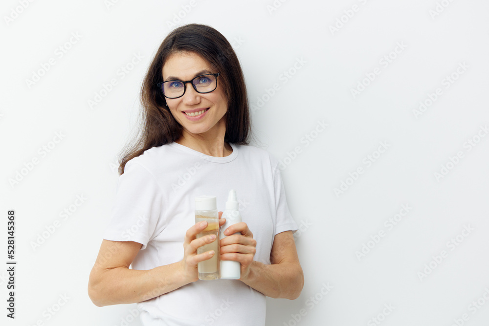 a funny brunette woman holds various jars of skin care cosmetics in her hands,standing in a white t-shirt and glasses on a light background with empty space a funny brunette woman holds various jars of skin care cosmetics in her hands,standing in a white t-shirt and glasses on a light background with empty space