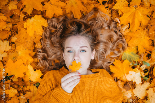 A beautiful young girl with blue eyes lies in the autumn leaves in the park and looks into the camera, closes her lips with a leaf.