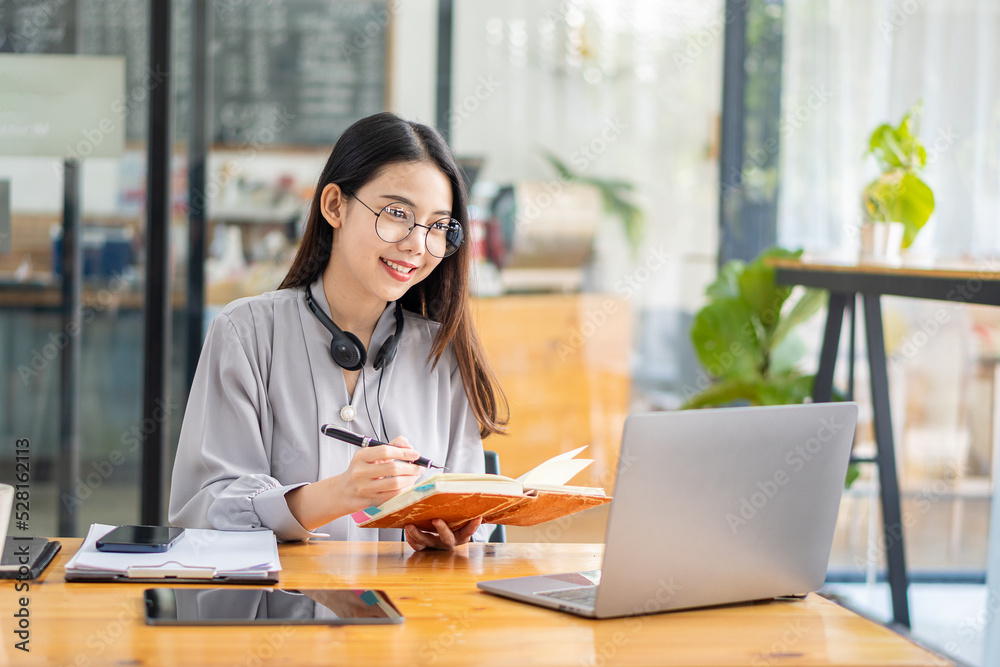 Pretty Asian woman and headphones using laptop in coffee shop writing notes. Attractive female student learning language. View webinars online. audio course education Pretty Asian woman and headphones using laptop in coffee shop writing notes. Attractive female student learning language. View webinars online. audio course education