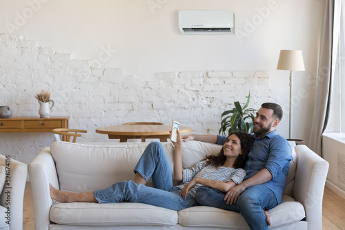 Couple enjoy fresh conditioned air inside cozy living room. Carefree wife holds remote controller adjust comfortable temperature relaxing on couch with husband at home. Tech for comfort life concept