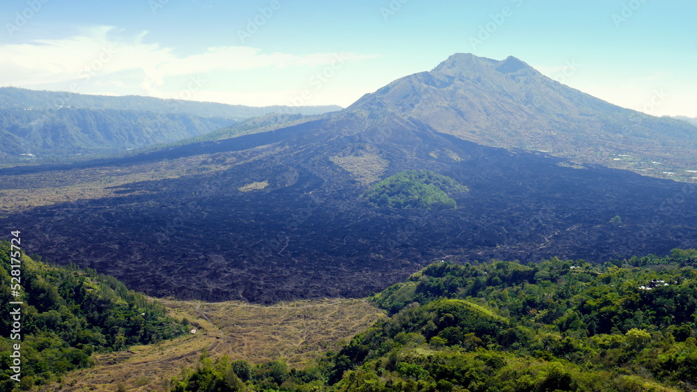 Vulkan Batur in Bali mit schwarzen Lavafeldern und grünem Wald unter blauem Himmel mit Nebel