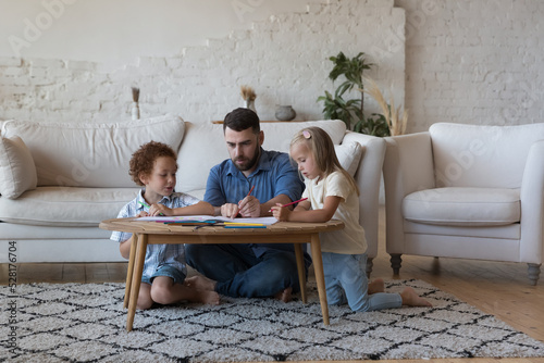 Loving father his diverse pre-schoolers children drawing pictures with colored pencils in sketchbook sit at table at home. Family weekend leisure, creative hobby and pastime, care and custody concept photo