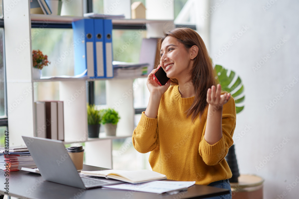 Obraz premium Beautiful Asian business woman chatting on the phone with customer and working on her laptop and taking note enthusiastically and happily in the office.