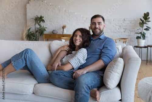 Multi ethnic millennial couple relaxing on comfortable couch smile pose for camera, spend free time, leisure at modern cosy home. Home-owners family portrait, bank loan for young family, love concept