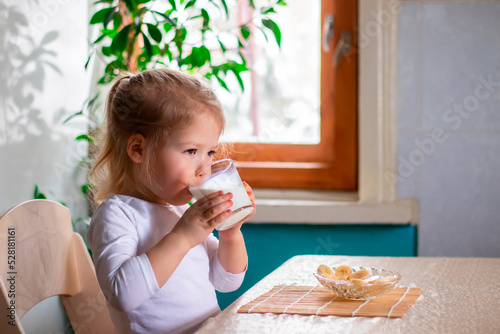 a little cheerful girl having breakfast with pieces of bananas and a glass of milk on the kitchen table in the morning. a child drinking a milk from a glass