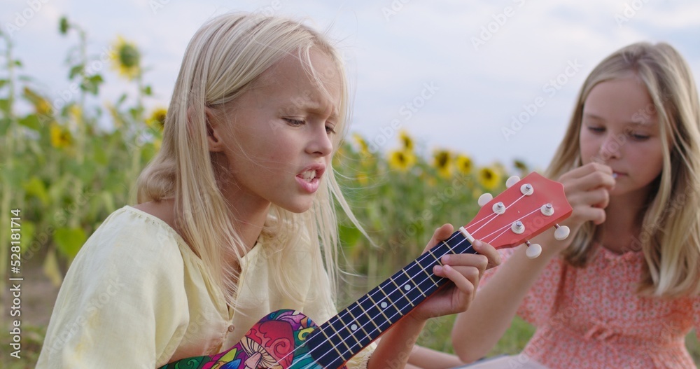 Fototapeta premium Portrait of joyful two girls sit on blanket in field, have picnic, read book, play ukulele guitar, eat sunflower seeds . Relaxing time. Happy sisters outdoors. Harvest. Childhood. Country life.