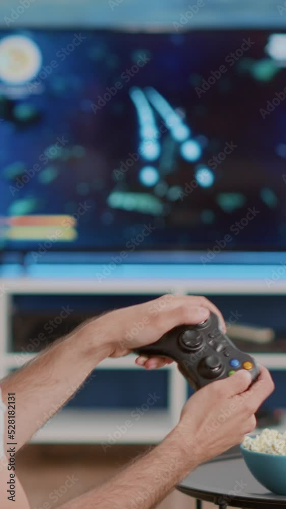 Man holding wireless controller playing console video game on tv while sitting on sofa in modern living room. Gamer relaxing on couch enjoying online gaming first person shooter on television.