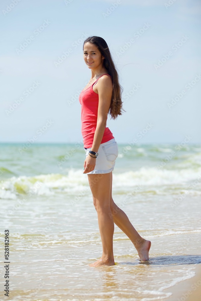 A young girl in red clothes and shorts stands on the beach or ocean with her feet in the water. Admiration for the sea surf and summer weather.