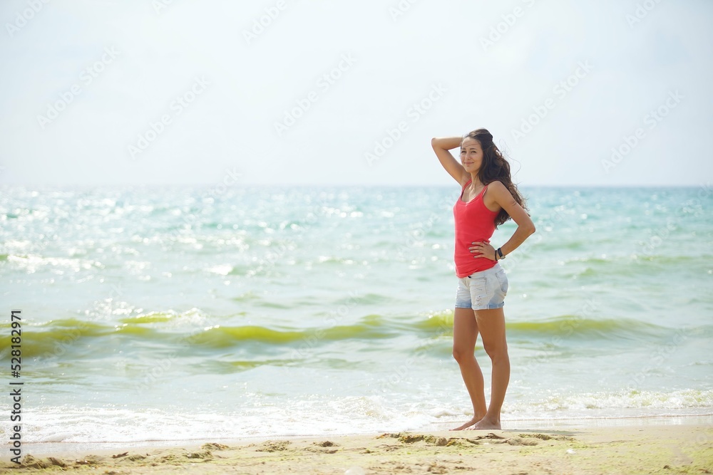 A young girl in red clothes and shorts stands on the beach or ocean with her feet in the water. Admiration for the sea surf and summer weather. A young girl in red clothes and shorts stands on the beach or ocean with her feet in the water. Admiration for the sea surf and summer weather.