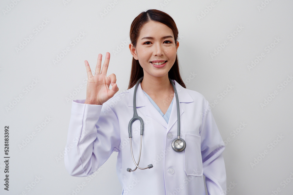 Happy smiling Asian female doctor wearing white medical uniform thumbs up ok with medical stethoscope on gray background medical concept. Happy smiling Asian female doctor wearing white medical uniform thumbs up ok with medical stethoscope on gray background medical concept.