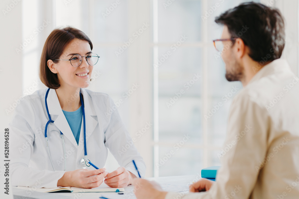 Photo of male patient writes all details makes notes during consultancy with doctor sit in spacious office. Physician tells treatment issues and explains diagnosis to hospital visitor. Medical concept Photo of male patient writes all details makes notes during consultancy with doctor sit in spacious office. Physician tells treatment issues and explains diagnosis to hospital visitor. Medical concept