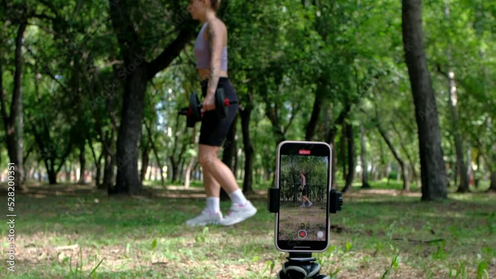 Beautiful blogger woman training in the park at summer day use smartphone for recording exercises or online lessons.