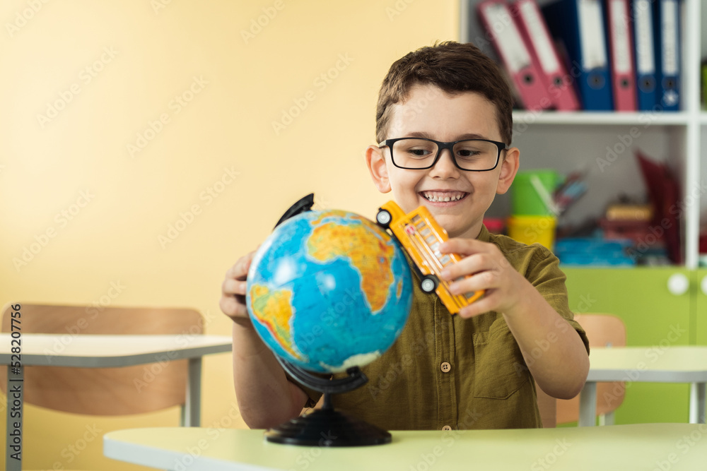 Cute caucasian schoolboy wearing glasses and sits at a desk in the classroom at elementary school. Little boy next the Globe playing with yellow school bus toy. Back to school concept Cute caucasian schoolboy wearing glasses and sits at a desk in the classroom at elementary school. Little boy next the Globe playing with yellow school bus toy. Back to school concept