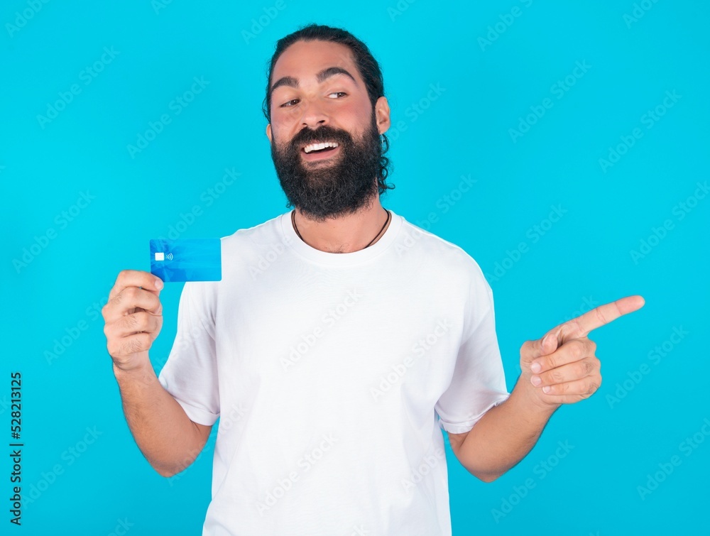 Curious smiling young bearded man wearing white T-shirt over blue studio background showing plastic bank showing finger copyspace Curious smiling young bearded man wearing white T-shirt over blue studio background showing plastic bank showing finger copyspace