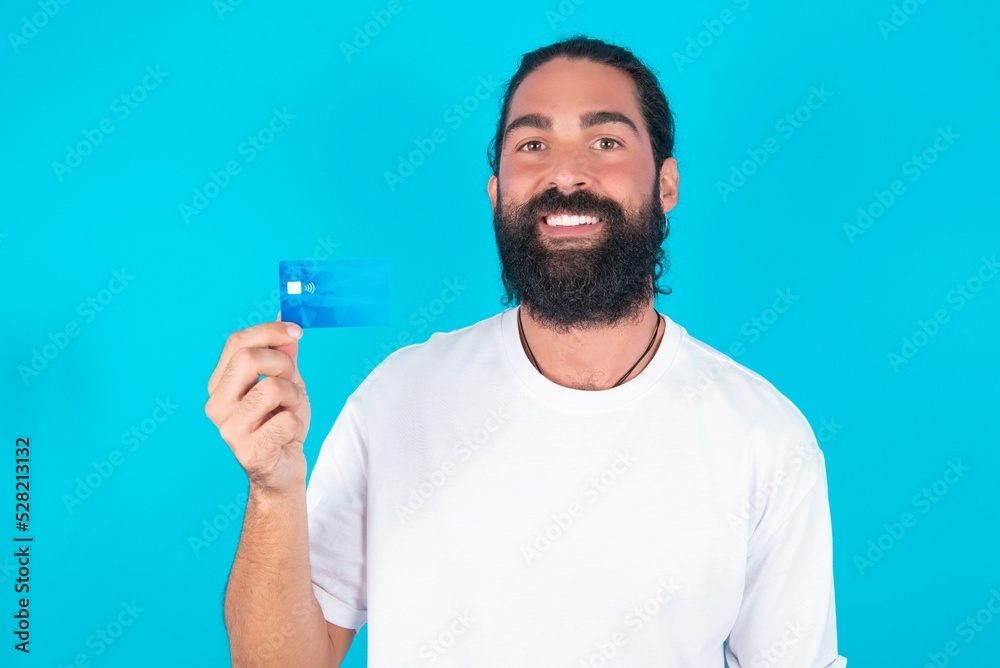 Photo of happy cheerful smiling positive young bearded man wearing white T-shirt over blue studio background recommend credit card Photo of happy cheerful smiling positive young bearded man wearing white T-shirt over blue studio background recommend credit card