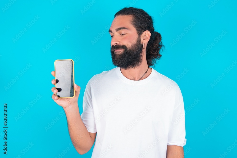 Photo of interested young bearded man wearing white T-shirt over blue studio background hold telephone look side empty space screen Photo of interested young bearded man wearing white T-shirt over blue studio background hold telephone look side empty space screen