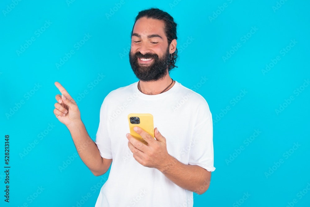 Smiling young bearded man wearing white T-shirt over blue studio background pointing finger at blank space holding phone in one hand Smiling young bearded man wearing white T-shirt over blue studio background pointing finger at blank space holding phone in one hand