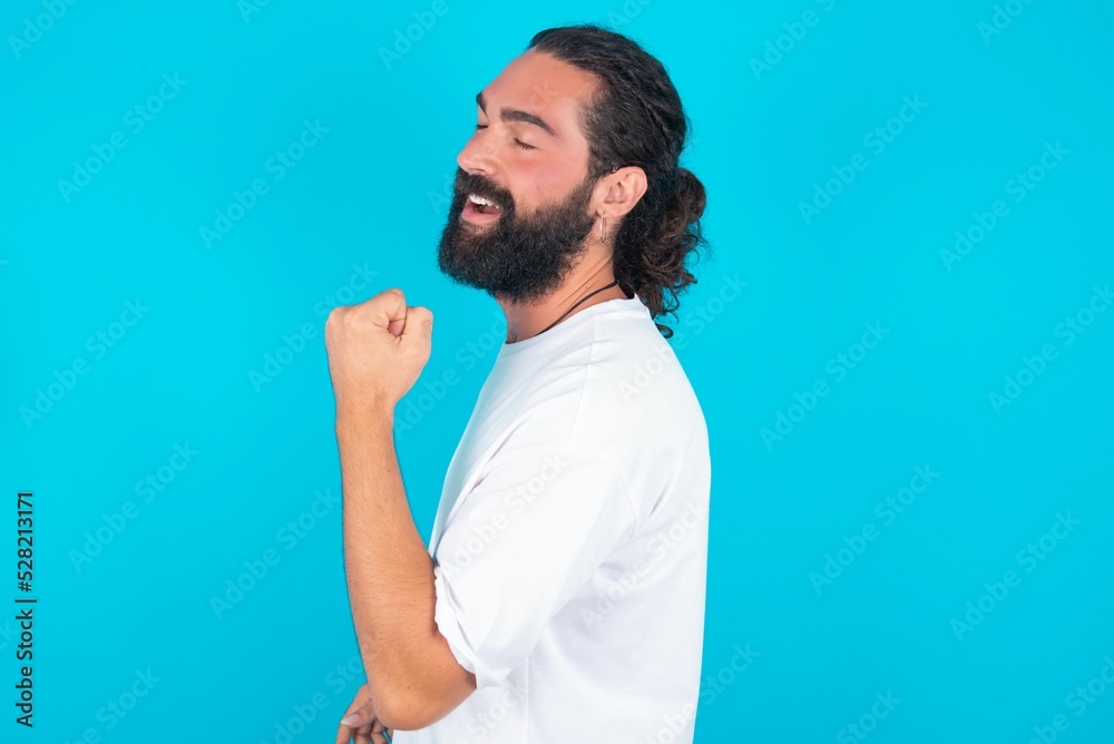 Profile side view portrait young bearded man wearing white T-shirt over blue studio background celebrates victory Profile side view portrait young bearded man wearing white T-shirt over blue studio background celebrates victory