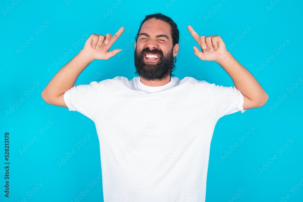 Photo of crazy young bearded man wearing white T-shirt over blue studio background screaming and pointing with fingers at hair closed eyes
