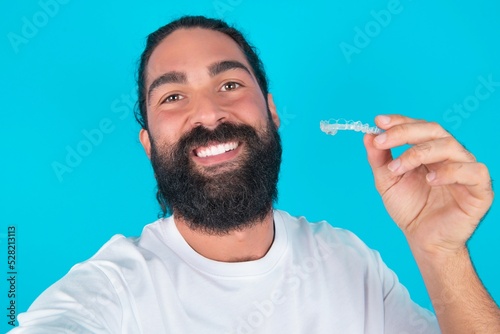 young bearded man wearing white T-shirt over blue studio background make selfie holding an invisible braces aligner, recommending this new treatment. Dental healthcare concept.