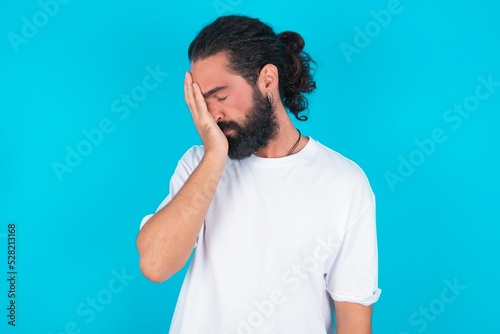 young bearded man wearing white T-shirt over blue studio background with sad expression covering face with hands while crying. Depression concept.