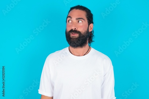 Shocked young bearded man wearing white T-shirt over blue studio background look empty space with open mouth screaming: Oh My God! I can't believe this.